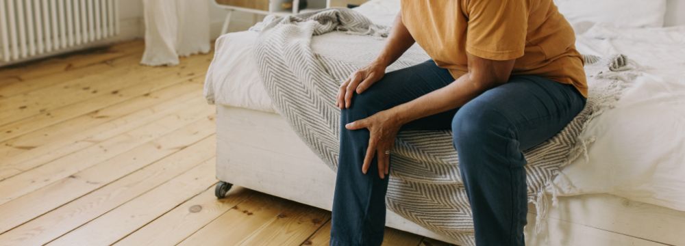 Woman sitting on edge of bed holding knee and leg in pain 