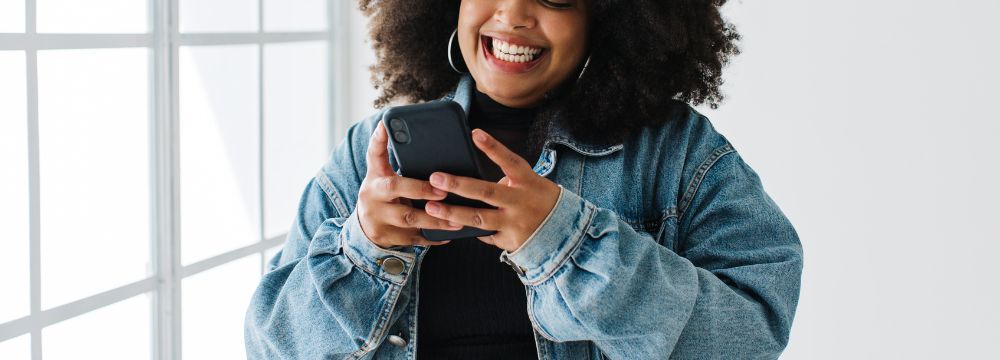 Woman next to window smiling while looking at phone and holding phone with both hands 